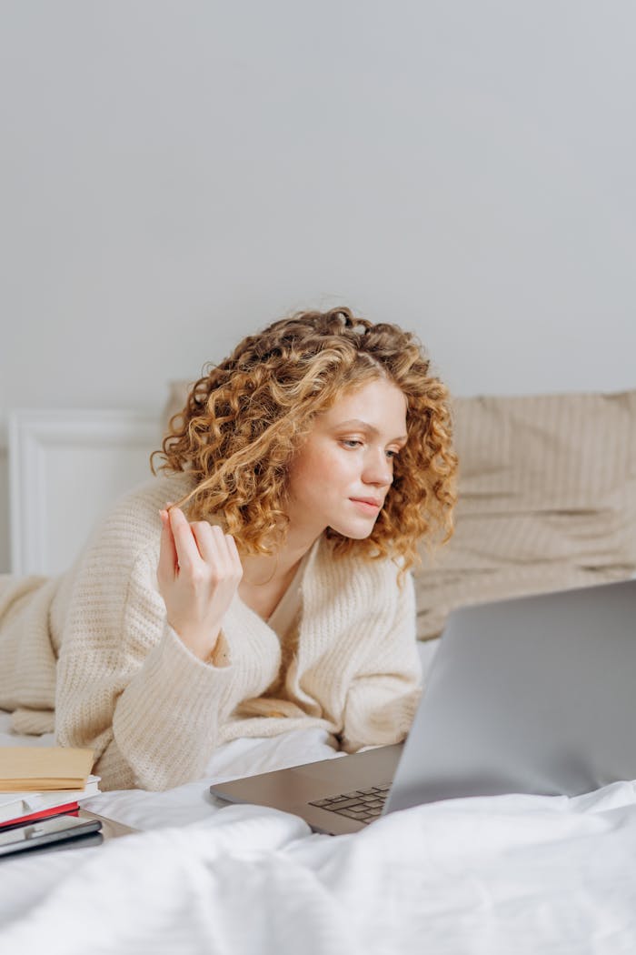 Caucasian woman with curly hair using a laptop while lying in bed, in a cozy home setting.